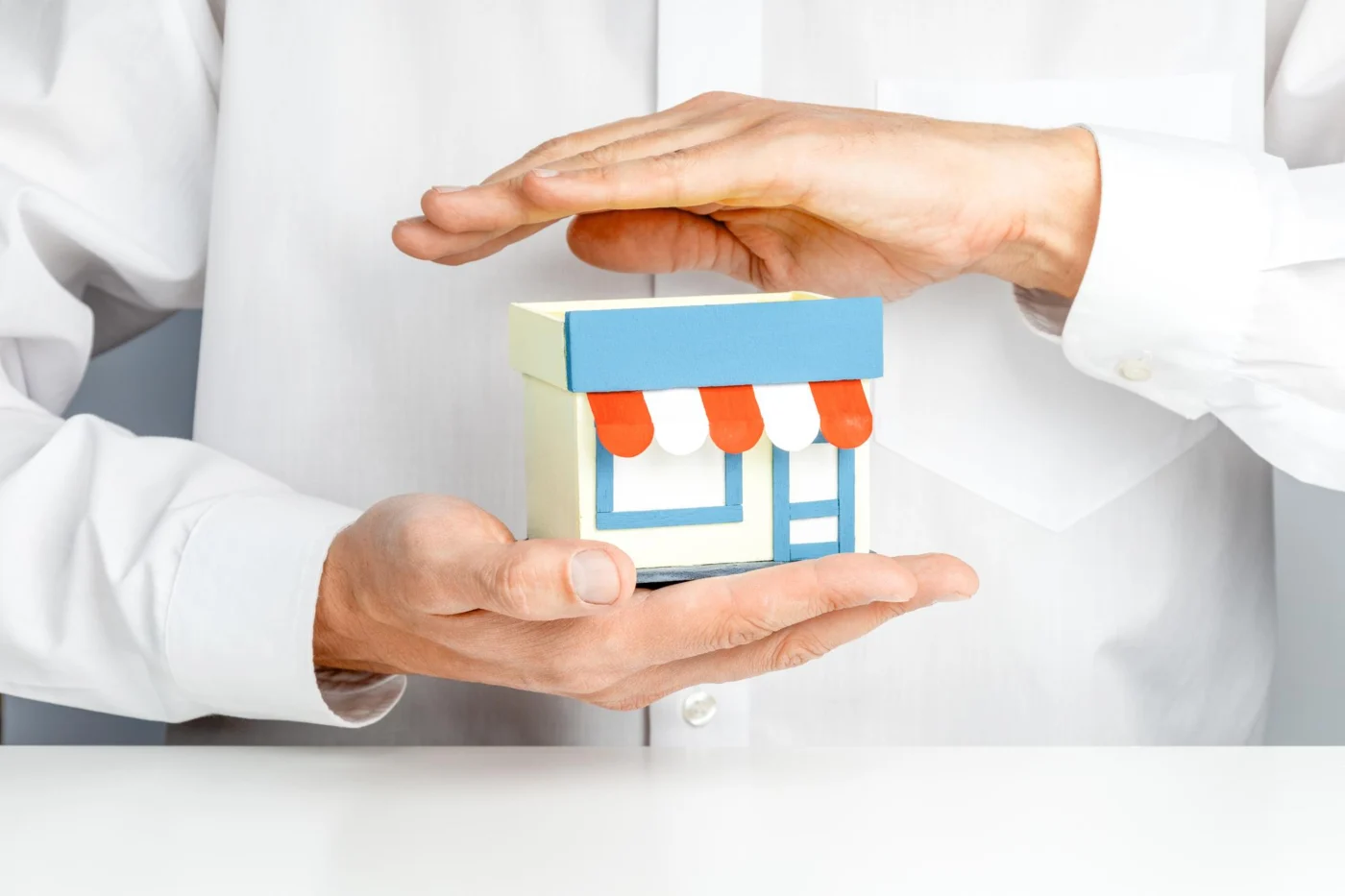 A pair of hands forming a protective shape over a small model storefront, representing small business protection