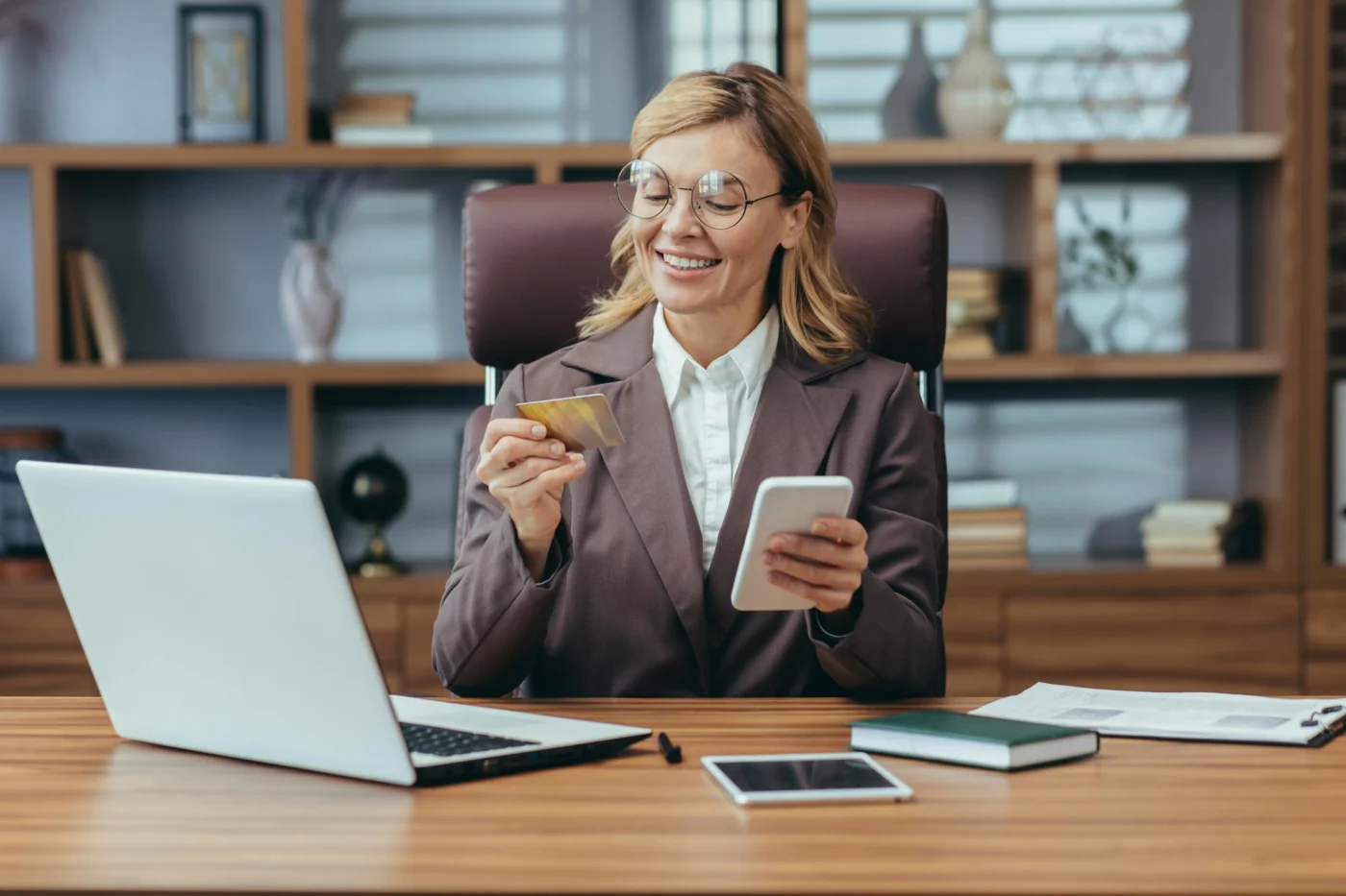 Smiling businesswoman at an office desk checking a smartphone next to her laptop, reviewing payment for freelance work